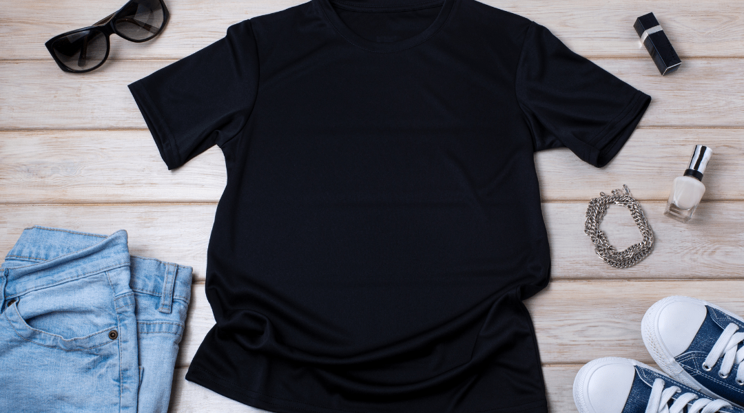 Flat lay of a black T-shirt styled with light blue jeans, white sneakers, nail polish, lipstick, sunglasses, and a silver chain bracelet on a wooden background.